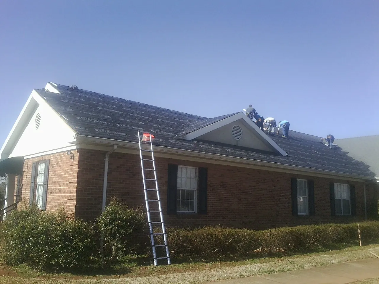 Crew tearing shingles off a brick ranch roof with a ladder set — Metal Roof Repair work by our crew in Cornish