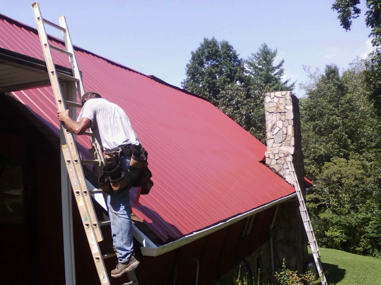 Roofer on a ladder beside a red metal roof and stone chimney — Soffit & Fascia Repair by Roofing Brothers in Cornish